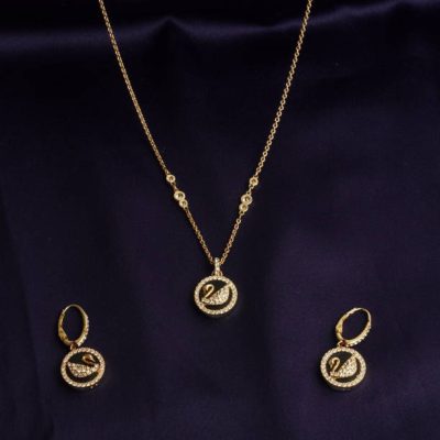 A set of one necklace and two earrings. Necklace has a gold chain and a pendant. The pendant and earrings are circular shaped with design of black and gold Swan. They have small white diamonds studded on the periphery. Close up on dark blue background.